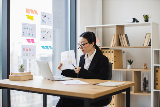 Confident Asian businesswoman wearing headset presenting data during video call using laptop. Middle-aged woman working remotely in office setting showcasing professional skills and communication. - Powered by Adobe