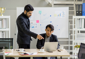 Two professional in suit engage in a discussion at a desk. One point at a laptop screen while the other attentively listen. Chart and graph on the wall provide data insight.