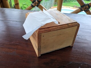 Wooden tissue box on a patio table in a tropical setting. A light-brown wooden tissue box, displaying a simple design, sits on a wooden table outdoors.