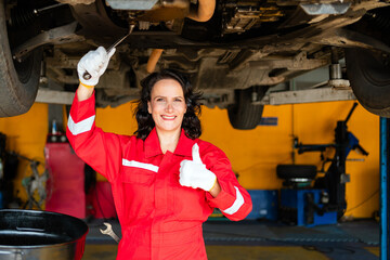A smiling professional female mechanic in red overalls works on a car's undercarriage, holding a wrench and giving a thumbs up in a bright garage setting. Car repair service. Vehicle maintenance