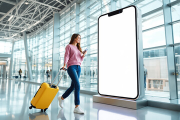 Woman walking in airport with luggage and smartphone beside a large screen.