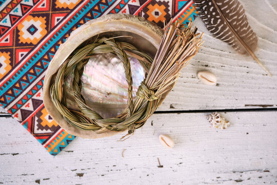Sweetgrass braid (Hierochloe odorata), also called vanilla grass, in an abalone shell with incense feather on fabric with native american pattern and old white wood. Overhead view.