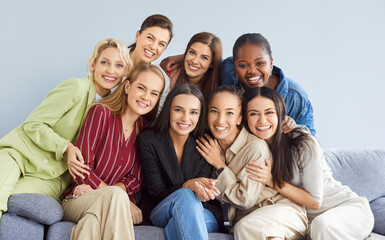 Portrait of group of female happy cheerful young diverse women friends coworkers or company employees hugging and looking at camera posing and smiling sitting on sofa on gray background.