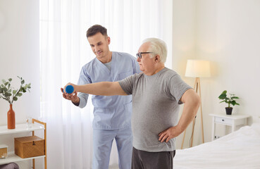 Fototapeta premium Chubby old pensioner man patient doing therapeutic rehab exercise with fitness dumbbell for arm muscles with assistance of young doctor physiotherapist or medic caregiver. Senior physiotherapy concept
