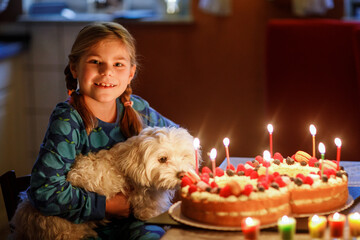 Happy little girl celebrating eighth birthday with her family Maltese dog. Cute smiling child with homemade cake as number eight, indoor. Happy healthy school kid blowing 8 candles on cake