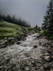 Misty Mountain River Winding Through Rugged Terrain In Foggy Weather With A Trist And Melancholic Atmosphere