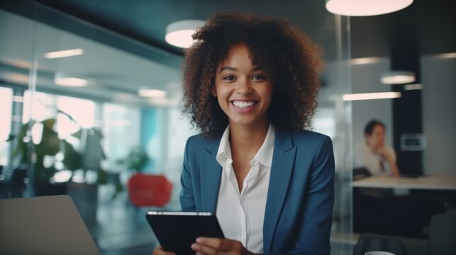 A cheerful young woman with curly hair uses a tablet in a bright, modern office. She wears a blue blazer and exudes confidence while sitting at a workspace