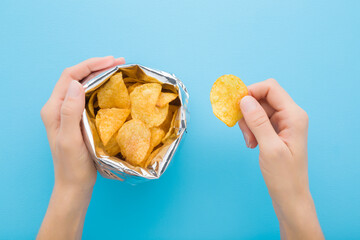 Young adult woman hands holding yellow potato chips and opened foil bag on light blue table...