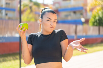 Young pretty brunette woman with an apple at outdoors making doubts gesture while lifting the shoulders