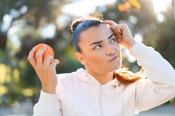 Young pretty brunette woman holding an orange at outdoors having doubts and with confuse face...