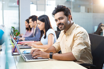 Young Professional Smiling At Camera While Typing On Laptop In Office