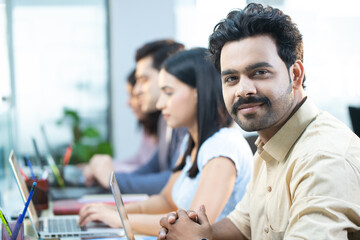 Work Colleague In Shirt Working On Laptop While Looking At Camera In Office