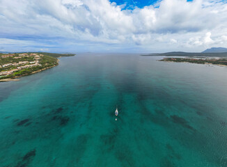 Aerial view of a serene turquoise sea in the north of Sardinia, Italy, with a lone sailboat in the center, surrounded by coastal greenery and distant horizons under a partly cloudy sky.