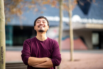 Portrait of a smiling Asian man looking up with arms crossed in an outdoor urban setting