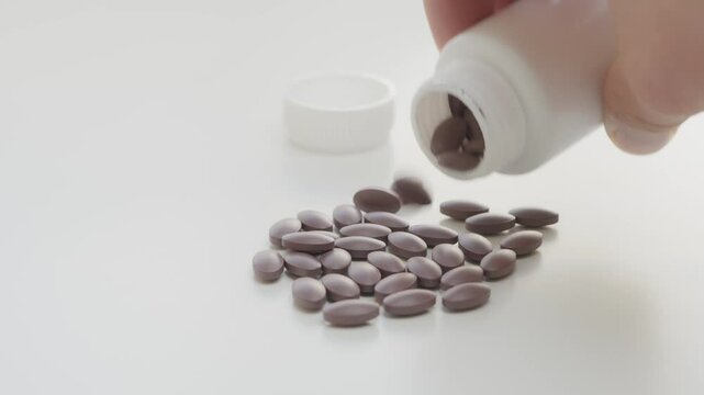 A white medicine bottle dispensing purple pills onto a white surface. A hand is tipping the bottle, emphasizing medication or health-related themes, and highlights pharmaceutical or healthcare concept