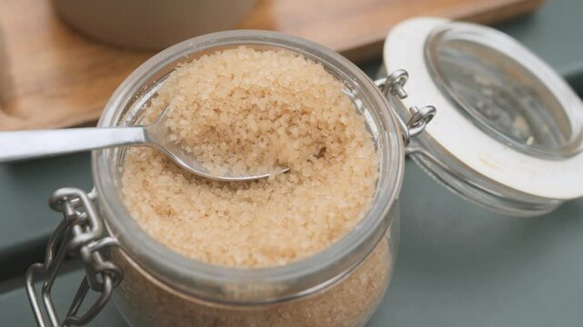 Close-up of an open glass jar containing granulated brown sugar with a metallic lid on a table.