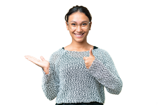 Young Uruguayan woman over isolated chroma key background holding copyspace imaginary on the palm to insert an ad and with thumbs up