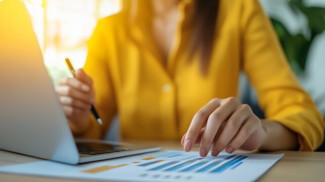 Woman working on laptop holding pen and writing down data from financial graph paper  - Powered by Adobe