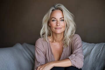 Obraz premium A serene middle-aged woman with flowing blonde hair sits comfortably on a soft couch, exuding a natural beauty and peaceful demeanor that represents confidence and self-assuredness.