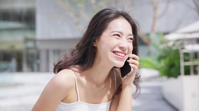 happy young asian woman talking on the mobile phone in the sunny urban street