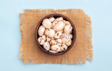 Top View of Fox Nut or Makhana in a Wooden Bowl on Burlap Fabric Isolated on Light Blue Background with Copy Space, Also Known as Lotus Seed Pop, Euryale Ferox or Gorgon Nut