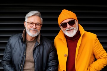Two fashionable older men exuding charm and style while posing for a photo, showcasing vibrant colors and smiles against a modern backdrop, celebrating their camaraderie.