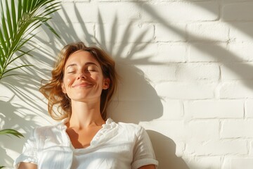 A serene woman relaxes against a white wall, soaking in sunlight and tranquility, surrounded by lush greenery, exemplifying inner peace and contentment amidst vibrant nature.
