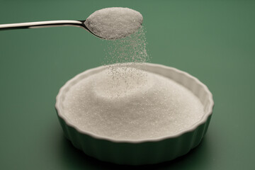 , Deutschland, 06.01.2025,  Ein Löffel mit rieselndem Erythrit über einer weißen Schale auf grünem Hintergrund. Darstellung einer natürlichen Zuckeralternative für bewusste Ernährung.