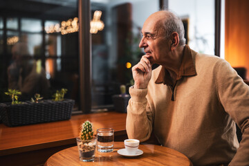 Sad senior man sitting by the window in the cafe and thinking about something while drinking coffee at evening.	