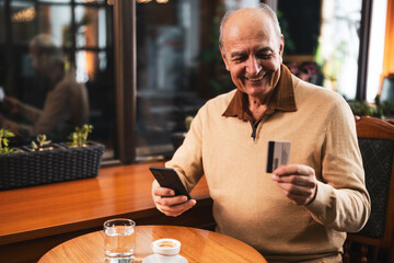 Smiling senior man in casual clothing using mobile phone and credit card while enjoying drinking coffee in the cozy city cafe.	