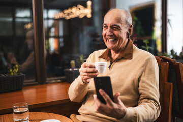 Smiling senior man drinking coffee while using mobile phone and earphones. He is relaxing in the cozy city bar at evening and using technology.	