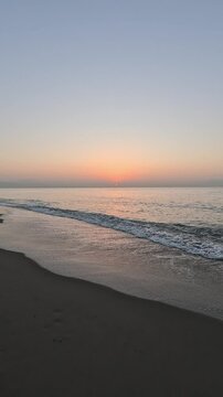 Vertical handheld video of a serene sunrise or sunset over a calm Mediterranean sea, with the sun casting a warm glow on the horizon at golden hour, viewed from the brown sand beach