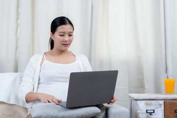Asian pregnant woman working on laptop while sitting on sofa in cozy home setting reflecting love, care, multitasking, maternal connection, and anticipation for future baby.
