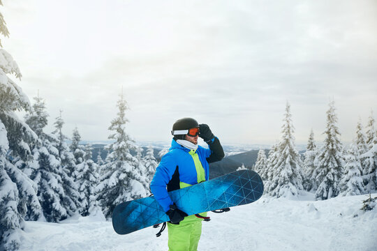 Man with snowboard standing amidst snowy landscape, surrounded by pine trees and a serene background.