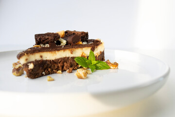 A piece of chocolate cake with nuts, decorated with a mint leaf, lies on a white plate. Small bowls with walnuts and peeled mandarin segments are located nearby. View from above