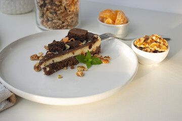 A piece of chocolate cake with nuts, decorated with a mint leaf, lies on a white plate. Small bowls with walnuts and peeled mandarin segments are located nearby. View from above