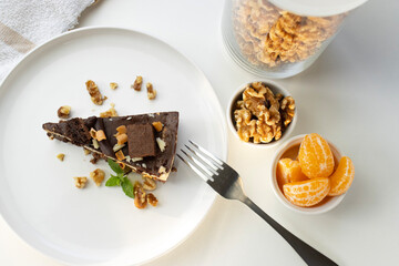 A piece of chocolate cake with nuts, decorated with a mint leaf, lies on a white plate. Small bowls with walnuts and peeled mandarin segments are located nearby. View from above
