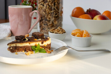 A piece of chocolate cake with nuts, decorated with a mint leaf, lies on a white plate. Small bowls with walnuts and peeled mandarin segments are located nearby. View from above