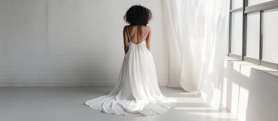 Elegant Woman with Natural Black Hair in White Boho Dress Posing in Minimalistic Light-Filled Studio with Large Window and Expansive Copyspace