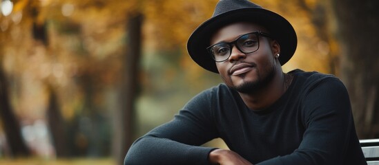 Obraz premium Stylish African man wearing a black hat and glasses, sitting on a park bench surrounded by autumn foliage, showcasing urban fashion in warm tones.