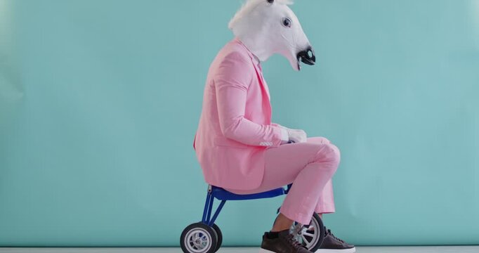 Funny humorous man riding tricycle like kid, wearing cute horse head mask, formal beautiful pink pastel color stylish suit, charming animal party show, strange comic character joke, playful racer
