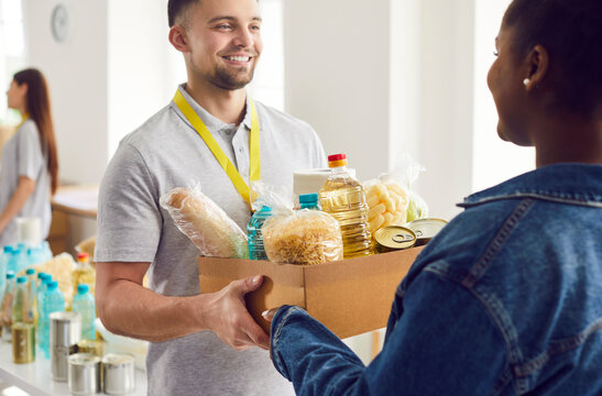Cheerful volunteer hands a box of food to an African-American refugee woman at a humanitarian aid center. The charity organization offers assistance, donations, and help to those in need.