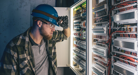 A male electrician works in a switchboard in overalls against the backdrop of emergency lighting.