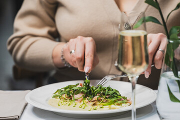 A close-up of a person enjoying a gourmet dish with greens and sauce, accompanied by a glass of sparkling beverage. The elegant dining atmosphere adds to the culinary experience.