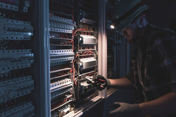 A male electrician works in a switchboard in overalls against the backdrop of emergency lighting.