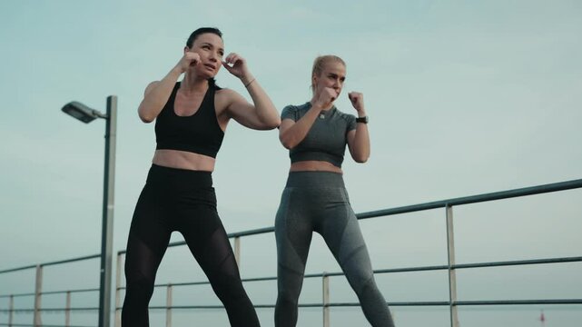 Two athletic women engage in kickboxing training outdoors, practicing leg kicks by the sea during sunset.