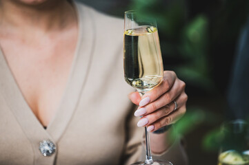 A close-up of a woman's hand elegantly holding a glass of sparkling wine. background features blurred greenery, creating a festive and sophisticated atmosphere. Perfect for celebrations or toasting.