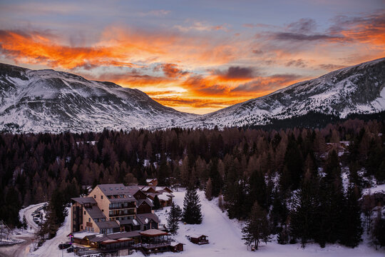 Vall&eacute;e enneig&eacute;e et bois&eacute;e entre deux montagnes avec village de station de ski sous un ciel orange nuageux du soleil levant en hiver.