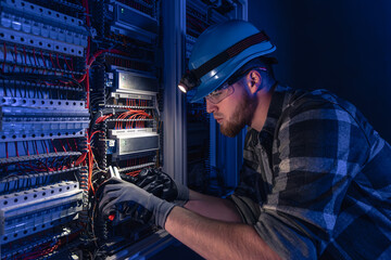 A male electrician works in a switchboard in overalls against the backdrop of emergency lighting.