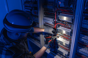A male electrician works in a switchboard in overalls against the backdrop of emergency lighting.